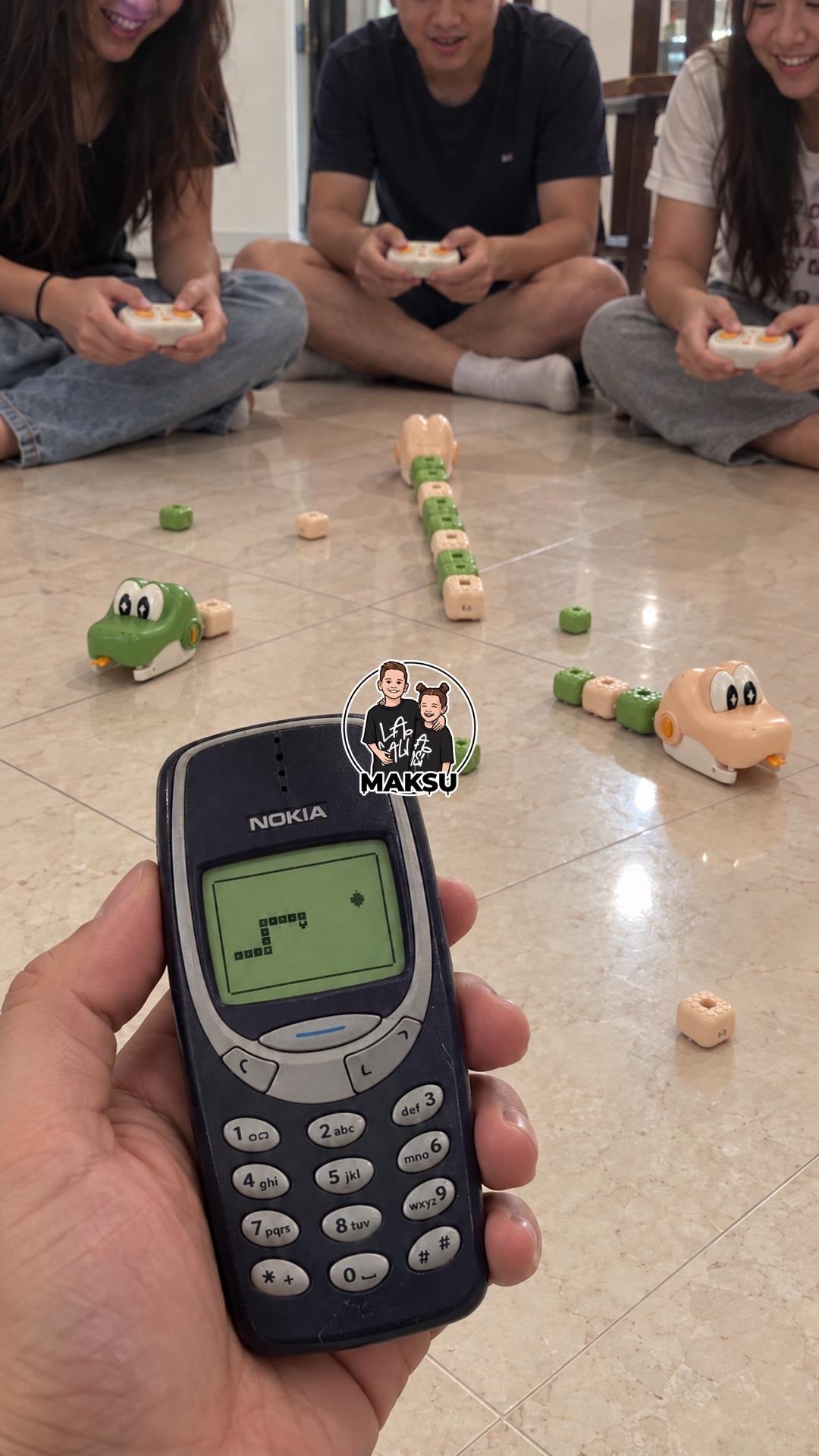 Three friends sitting on the floor racing remote control magnetic snakes while a hand holds an old Nokia phone with the Snake game